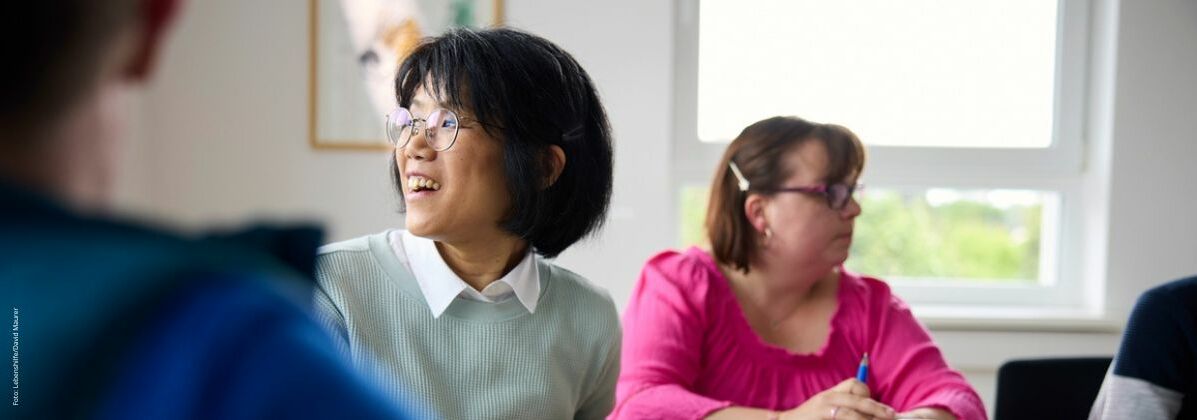 Zwei lächelnde Frauen sitzen mit anderen an einem Besprechungstisch. Im Arbeitsalltag bei der Lebenshilfe Würzburg steht der Mensch im Fokus.