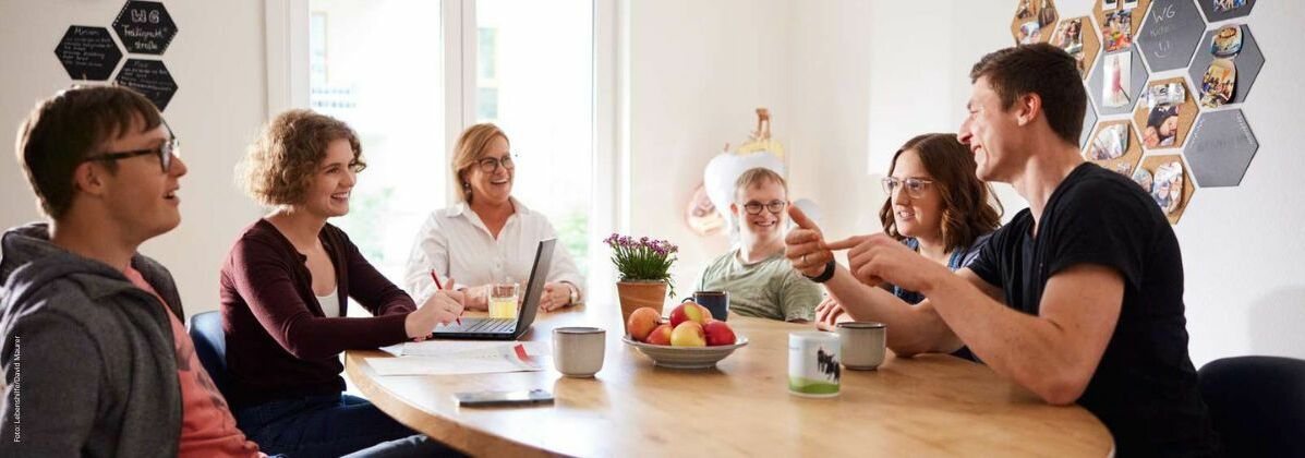 Menschen mit und ohne Behinderung sitzen gemeinsam am Tisch. Die Lebenshilfe Würzburg e.V. ermöglicht in ihren inklusiven Wohngemeinschaften selbstbestimmtes und gemeinschaftliches Wohnen.