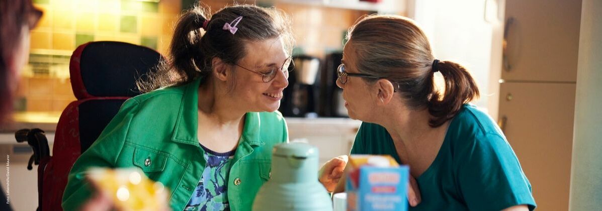 Zwei lächelnde Frauen mit und ohne Behinderung sitzen gemeinsam am Frühstückstisch.